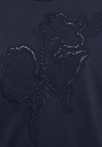 Camicia blu navy con intricati ricami floreali blu scuro, con dettagli in rilievo e una finitura testurizzata. Tessuto liscio con collo rotondo.