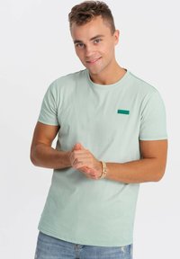 Světlezelené tričko vyrobené z bavlny, kulatý výstřih, krátké rukávy, má malý zelený obdélníkový nášivku na hrudi.