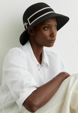 Tume nahaga naine, kes kannab musta laiade äärega mütsi valge ribaga ja valget pluusi, istub valge seina ääres ning vaatab küljele.