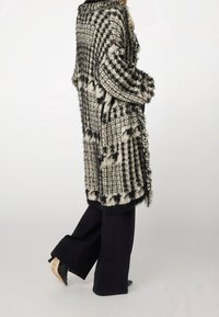 Persona che indossa un cardigan a maglia con frange nero e grigio sopra pantaloni neri a gamba larga e tacchi a punta neri, in posizione laterale.