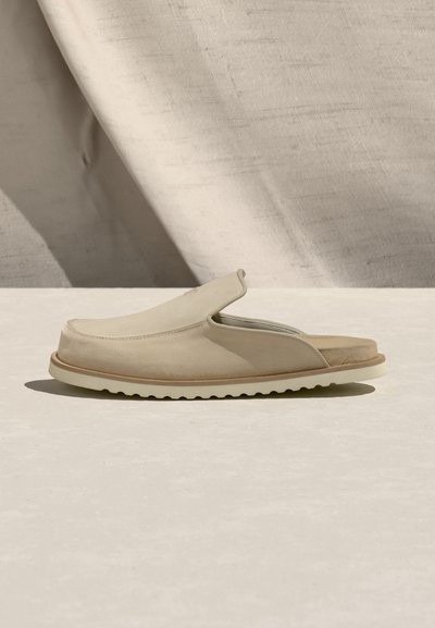 Μπεζ δερμάτινο mule χωρίς κορδόνια με κλειστή στρογγυλεμένη μύτη, ανοιχτή φτέρνα και λευκή ραβδωτή σόλα σε ανοιχτό μπεζ επιφάνεια και υφασμάτινο φόντο.