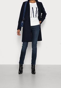 Cappotto blu sopra una camicia bianca con stampa "GAP", abbinato a jeans scuri e stivaletti neri alla caviglia. Presenta una borsa a tracolla blu.