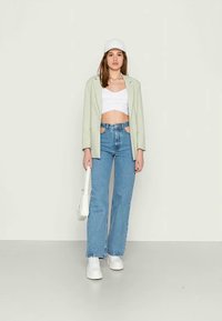 Blazer verde claro, top curto branco, jeans azuis de cintura alta, sapatilhas brancas e um boné branco. Modelo segurando uma pequena bolsa branca.
