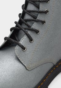 Gros plan d'une botte en cuir gris avec des lacets noirs mouchetés passés dans des œillets en métal et des détails de couture jaune sur la semelle.