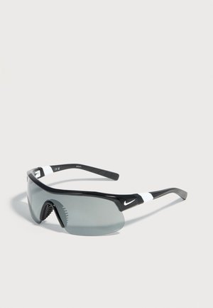 Schwarz-weiße Nike-Sportsonnenbrille mit gebogenen grauen Gläsern, Gumminasenpads und flexiblen Bügeln auf einer grauen Oberfläche.