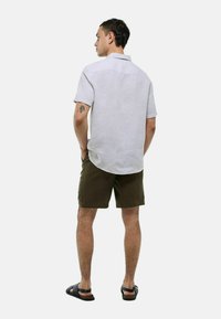 Chemise à manches courtes boutonnée gris clair avec col, associée à un short vert olive et des sandales noires, illustrant un look estival décontracté.