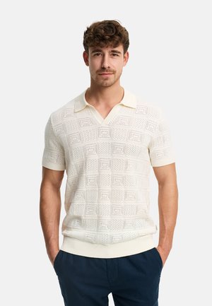 Mężczyzna o krótkich kręconych włosach, ubrany w kremową, krótkorękawową dzianinową koszulkę polo z teksturowanym wzorem w kwadraty oraz granatowe spodnie, z rękami w kieszeniach.