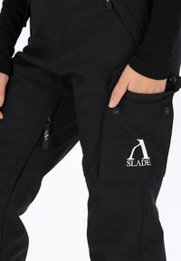 Pantaloni softshell neri con tasca laterale e logo, caratterizzati da diverse texture, design aderente, cerniere e dettagli in metallo.