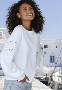 Felpa bianca con intricati disegni ritagliati sulle maniche, abbinata a jeans in denim azzurro chiaro, su uno sfondo luminoso.