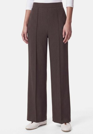 Pantalon marron à jambes larges avec fines rayures blanches verticales, porté avec des chaussures blanches et un haut blanc à manches longues.