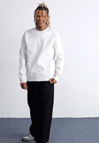 Hvid bomulds sweatshirt med rund hals, ribbede manchetter og et guldaccent. Løst siddende sorte bukser. Grå sneakers. Ensfarvet baggrund.