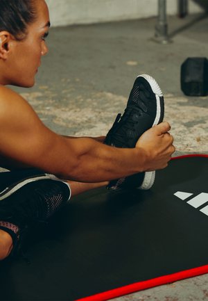 Schwarze Sportschuhe mit Netzakzenten und Gummisohlen. Die Person dehnt sich auf einer schwarzen Gymnastikmatte mit rotem Rand und weißem Logo.