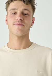 Beige cotton t-shirt featuring a crew neck and a small embroidered logo on the left side. Smooth texture and relaxed fit.