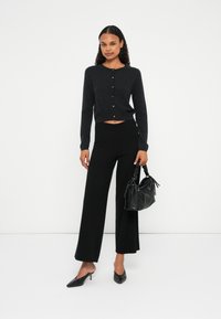 Μαύρος κοντός κα cardigan με κουμπιά, συνδυασμένος με φαρδιά μαύρη παντελόνα. Το μοντέλο κρατάει μια μικρή μαύρη τσάντα και φοράει μυτερές μαύρες γόβες.