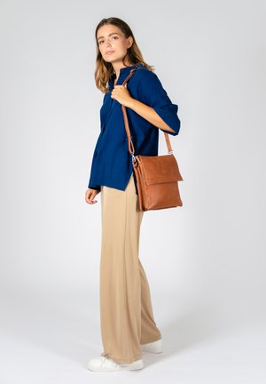 Sac en cuir marron avec rabat avant, bandoulière ajustable et texture lisse. Assorti à une chemise bleue à boutons et un pantalon beige.