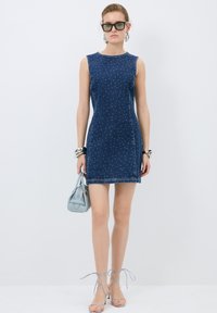 Denim sleeveless dress with a fitted design, blue with white polka dots, and visible stitching. Paired with strappy sandals and a small handbag.
