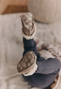 Brązowe kapcie dziecięce z białą, futrzaną wyściółką i wzorem misia na podeszwach, zestawione z ciemnoniebieskimi legginsami z teksturowanej tkaniny.