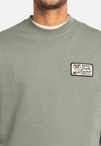Groene sweatshirt met een gestructureerd oppervlak, voorzien van een zwart-wit slang controle patch op de linkerborst. Klassieke ronde hals.