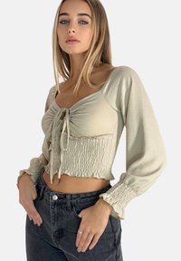 Ljusbeige kort topp med knytning framtill, smockat midja och långa puffärmar. Texturerad tyg med en mjuk yta. Matchad med mörka jeans.