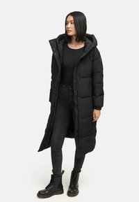 Sort lang pufferjakke med hætte, featuring en lynlås lukning og quiltet design. Båret over en sort top og kombineret med mørke jeans og sorte støvler.