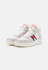 Tenisi high-top fabricați din piele bej și albă cu model, având accente roșii și negre, cu o talpă texturată și un logo proeminent.