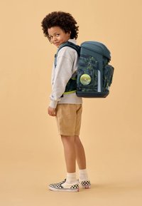 Dunkelblauer Rucksack mit grünem Spritzdesign, verstellbaren Trägern und mehreren Fächern, getragen von einem Kind in einem hellgrauen Sweatshirt und beigen Shorts.