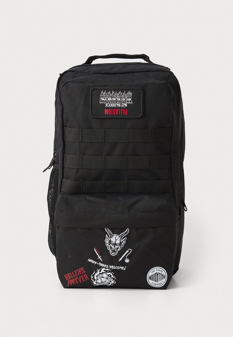Mochila de lona preta com um grande emblema, múltiplas alças e vários gráficos bordados na parte frontal, incluindo um diabo estilo cartoon e texto.
