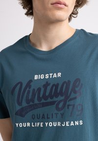 Tirkīza kokvilnas t-krekls ar ekrāndrukātām uzrakstām "BIG STAR Vintage Quality 79" tumši zilā un baltā krāsā. Klasisks apaļā kakla dizains.