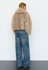 Vrouw zijwaarts kijkend, gekleed in een cropped beige bontjas en wijd uitlopende blauwe jeans met witte verfspatten, staand tegen een witte muur.