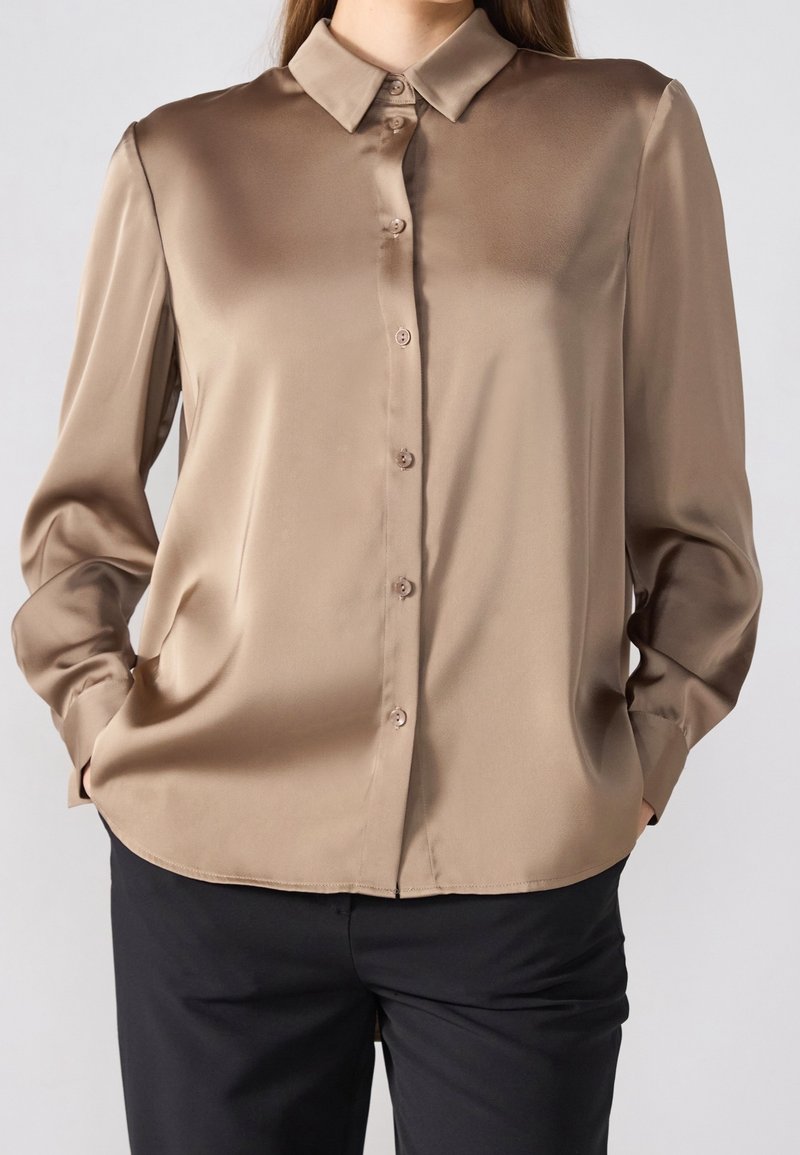 Femme portant un chemisier en satin taupe à manches longues boutonné, associé à un pantalon noir, mains dans les poches, sur un fond uni.