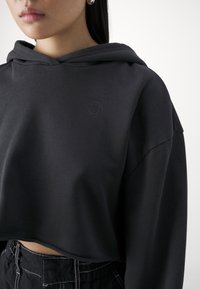 Sweat à capuche noir raccourci avec une texture douce, doté d'une capuche et d'un logo discret. Associé à un pantalon taille haute en tissu sombre avec des coutures claires.