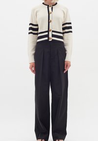 Krämfärgad kofta med svarta ränder, knappar framtill. I kombination med höga svarta byxor, med veck och en slät textur.