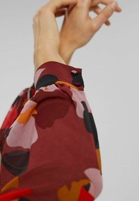 Fluffy blus med en vinröd bas, med stora blommönster i rosa, orange och svart. Volangärmsmanschett med knapprad.