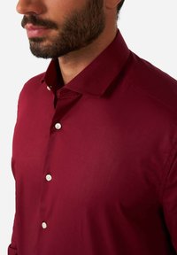 Camisa burdeos de botones con cuello estructurado, que presenta botones blancos y una tela suave y texturizada. Vista de cerca del escote y el hombro.