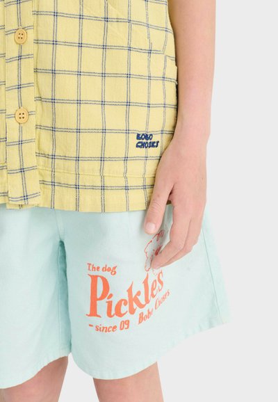 Main d'enfant reposant à côté d'un short bleu clair avec le texte orange "Pickles" et d'une chemise à carreaux jaune clair avec un petit texte bleu sur la poche.