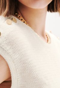 Gehaakt crèmekleurig sweater met textuurstof, ronde halslijn en gouden decoratieve knopen op de schouder. Gouden kettinghalsketting zichtbaar.