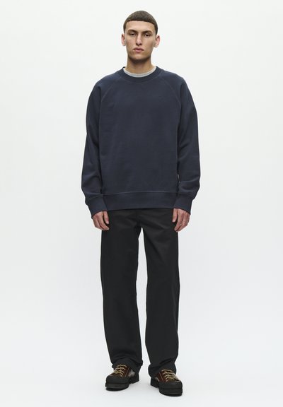 Mladi moški, oblečen v mornarsko modro majico s krožnim vratom, črne hlače in temno rjave čipkaste čevlje, stoji na belem ozadju.