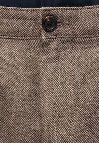 Tissu marron à motif chevrons avec un bouton noir à quatre trous attaché à la ceinture d'un vêtement.