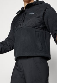Chaqueta negra corta que presenta una sección superior acolchada y una sección inferior de forro polar. Diseñada con capucha y bolsillo lateral para mayor funcionalidad.