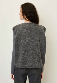 Pelēka fliša veste ar teksturētu, pūkainu virsmu, bez piedurknēm un noapaļotu apakšmalu. Uzvilkta virs garām piedurknēm piederīgas, pieguļošas augšas un pelēkjām biksēm.