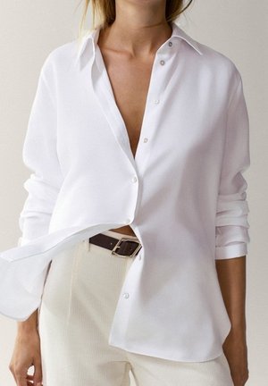 Femme portant une chemise blanche oversized à boutons avec manches retroussées, un pantalon beige et une ceinture noire avec une boucle argentée.