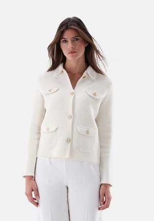 Weißer, strukturierter Cardigan mit kragenförmigem Ausschnitt, vier Taschen und goldenen Knopfakzenten. Kombiniert mit cremefarbenen Hosen.