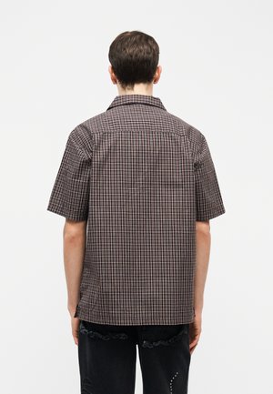 Mann trägt braun-rot karierte kurzärmelige Hemd und schwarze zerrissene Jeans, steht mit dem Rücken zum Betrachter vor einfachem Hintergrund.