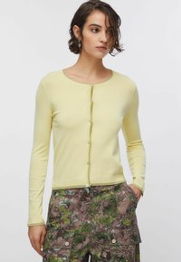 Sárga kardigán hosszú ujjakkal, kerek nyakkivágással és gombokból álló sorral. Kontrasztos szegéllyel és puha kötött textúrával rendelkezik.