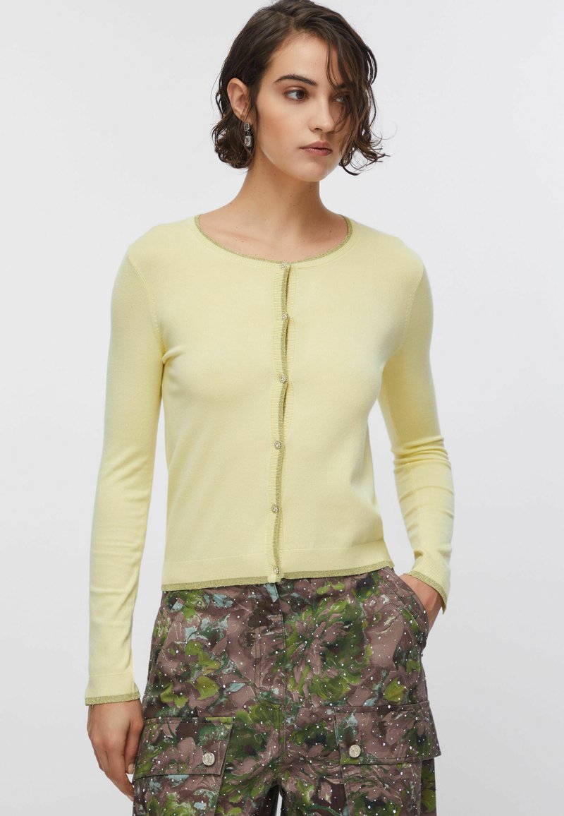 Sárga kardigán hosszú ujjakkal, kerek nyakkivágással és gombokból álló sorral. Kontrasztos szegéllyel és puha kötött textúrával rendelkezik.