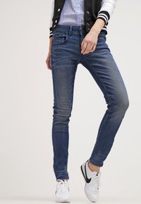 Vaqueros skinny de mezclilla azul con un lavado desgastado, cinco bolsillos y cierre de botón frontal, combinados con zapatillas blancas.