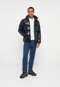 Man wearing a black puffer jacket over a white shirt, blue jeans, and black sneakers, standing against a white background.