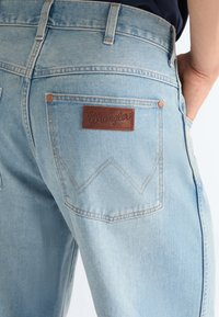 Gaiši zili džinsa džinsi ar Wranglera ādas etiķeti aizmugurējā kabatā, valkāti personas, kurai roka atbalstīta priekšējā kabatā.