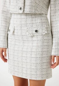 Witte en grijze tweed rok met een gestructureerde pasvorm, voorzien van twee voorzakken met zwarte knopen en een textuurpatroon op de stof.