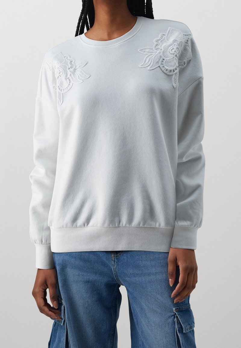 Personne portant un sweat-shirt blanc avec des détails en dentelle florale sur les épaules et un jean cargo bleu, debout devant un fond uni.